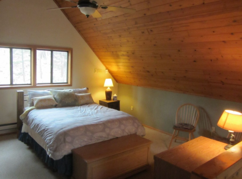 Cozy bedroom with a sloped wooden ceiling, a bed, two lamps, and a chair, surrounded by natural light from windows.