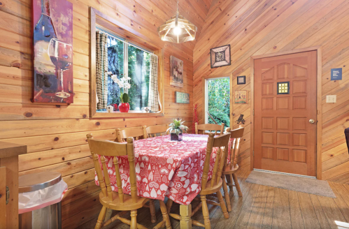 Cozy wooden dining area with a red tablecloth, surrounded by chairs and a window overlooking greenery.