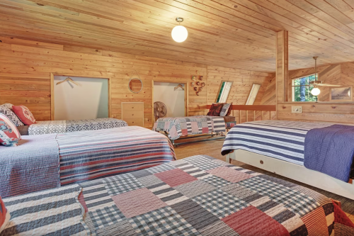 Cozy wooden attic bedroom with three beds, patterned quilts, and large windows for natural light.