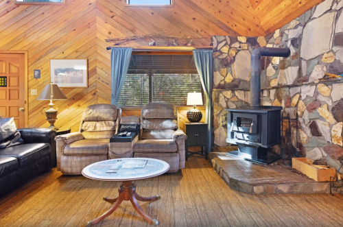 Cozy living room with wooden walls, a stone fireplace, two recliners, and a round coffee table. Warm and inviting atmosphere.