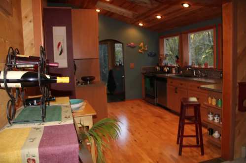 Cozy kitchen with wooden accents, wine rack, and large windows overlooking a forested area. Warm, inviting atmosphere.