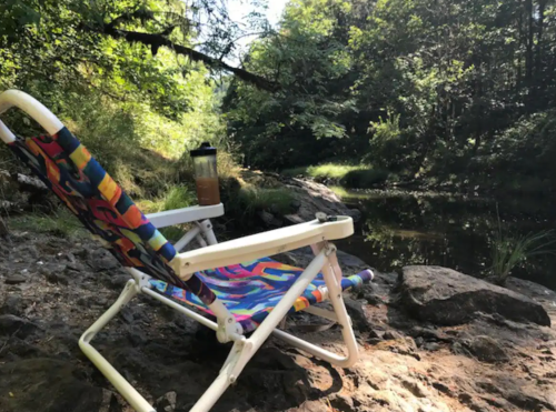 A colorful lounge chair by a serene riverbank, surrounded by trees and rocks, with a drink nearby.