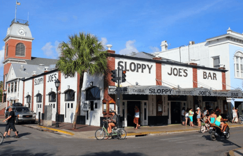 Exterior of Sloppy Joe's Bar in Key West, with people walking and biking outside under a clear blue sky.