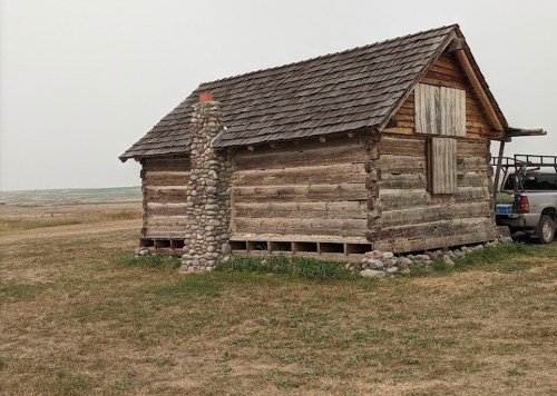 A rustic wooden cabin with a stone chimney, set in a grassy field under a hazy sky.