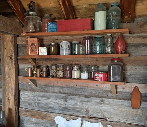 Wooden shelves in a rustic setting display various jars, containers, and a red book against a weathered wall.