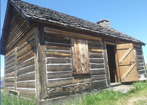 A rustic wooden cabin with a sloped roof, partially boarded windows, and a large open door, set against a clear blue sky.
