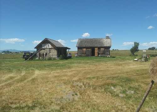 Two rustic wooden buildings on a grassy field under a clear blue sky, with distant hills in the background.