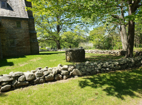 A stone well surrounded by grass and trees, with a historic building in the background on a sunny day.