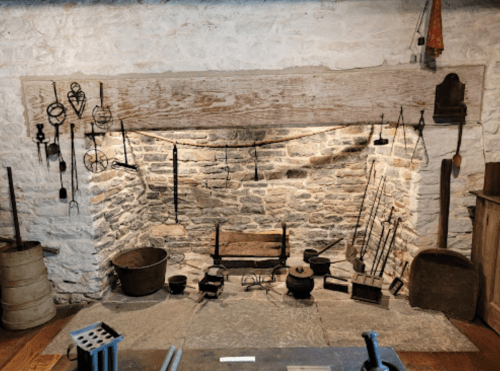 A rustic stone fireplace with various old cooking tools and utensils displayed on the wall and floor.
