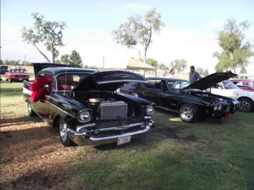 Two classic cars parked on grass, one black 1957 Chevrolet with the hood open and another black muscle car beside it.