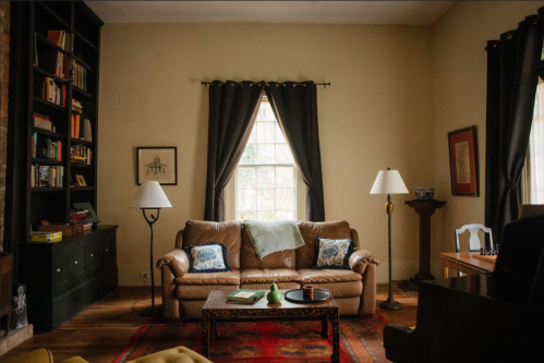 Cozy living room with a brown sofa, bookshelves, lamps, and a window draped in dark curtains. Warm, inviting atmosphere.