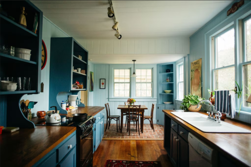 Bright kitchen with blue walls, wooden countertops, and a dining table, featuring large windows and plants.
