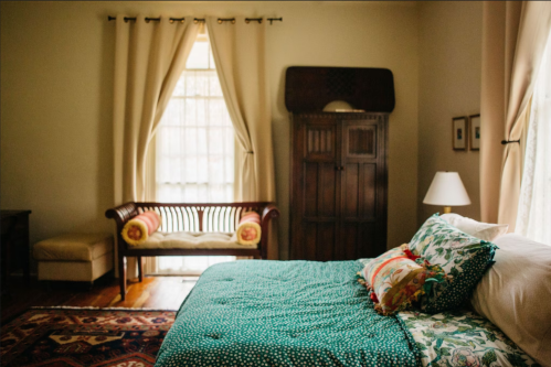 Cozy bedroom with a green quilt, decorative pillows, a wooden chair, and soft curtains letting in natural light.