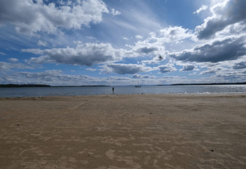 A serene beach scene with a person standing near the water under a cloudy sky, with a boat in the distance.