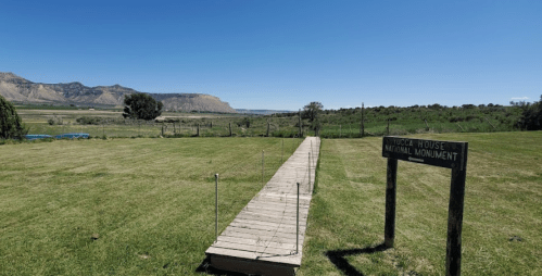 A wooden walkway leads to a sign for the Hovenweep National Monument, with a vast landscape and blue sky in the background.