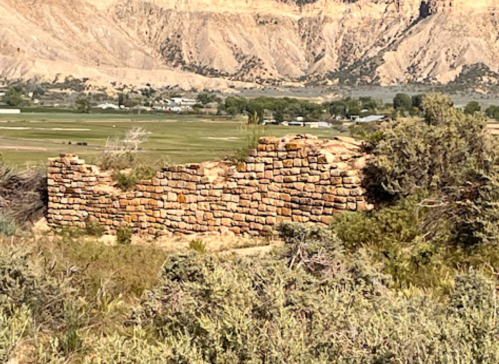 Ruins of a stone wall amidst grassy terrain, with mountains and fields in the background.
