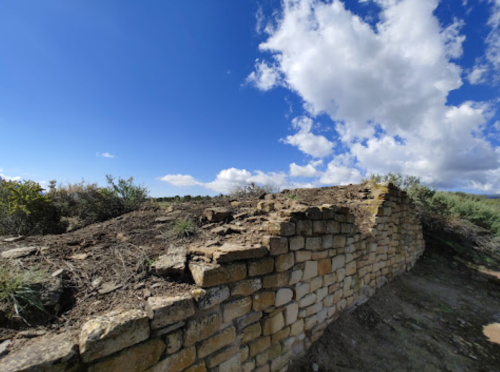 Ruins of a stone wall on a hillside under a blue sky with scattered clouds and surrounding vegetation.