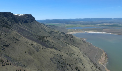 Aerial view of a rugged mountain landscape with green slopes, a lake, and distant hills under a clear blue sky.