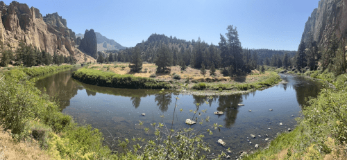 A serene river curves through a lush landscape, surrounded by rocky cliffs and pine trees under a clear blue sky.