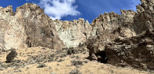 Rocky cliffs rise against a blue sky, with dry grass and sparse vegetation in the foreground.