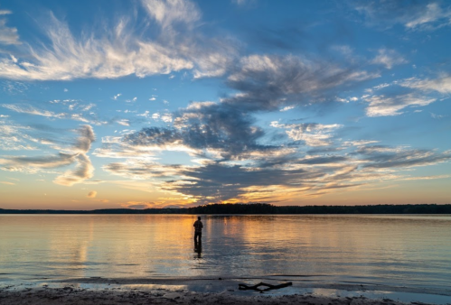 A person stands in calm water at sunset, with vibrant clouds reflecting in the sky and water.