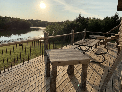 A wooden deck with tables overlooks a calm lake at sunset, surrounded by trees and a clear sky.