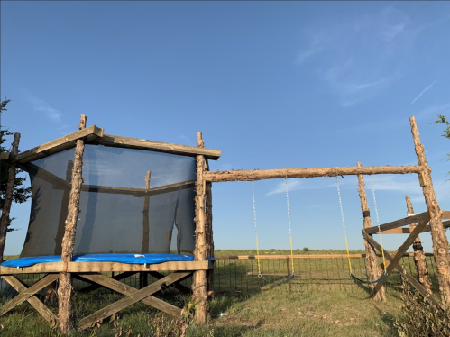 A wooden play structure with a trampoline and swings, set against a clear blue sky and open field.