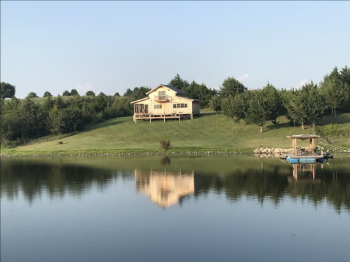 A yellow house on a hill reflects in a calm lake, surrounded by trees and a small dock.