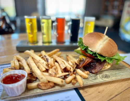 A delicious burger with lettuce, tomato, and bacon, served with fries and a side of ketchup, alongside three colorful beer samples.