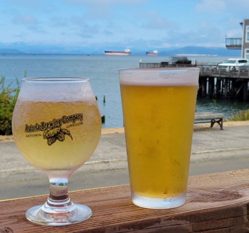 Two glasses of beer sit on a wooden railing, with a scenic view of the water and ships in the background.