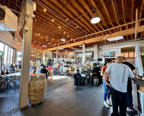 A lively brewery interior with wooden beams, string lights, and patrons enjoying drinks at tables and the bar.