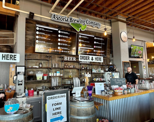Interior of Astoria Brewing Company with a bar, menu screens, and a staff member at the counter.