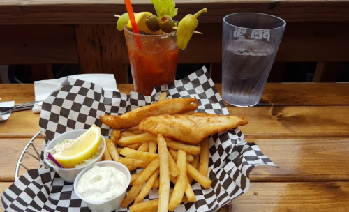 A plate of fried fish and fries with lemon, tartar sauce, and a Bloody Mary with olives on a wooden table.