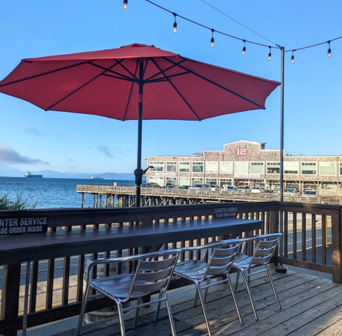 A red umbrella shades empty metal chairs on a wooden deck overlooking the water and a building in the background.