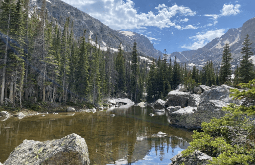 A serene mountain landscape with a clear river, rocky banks, and tall pine trees under a blue sky with scattered clouds.