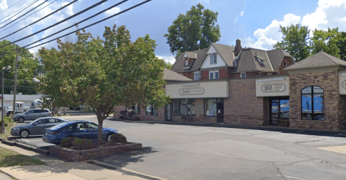 A street view of a brick building with shops, a tree, and parked cars under a partly cloudy sky.