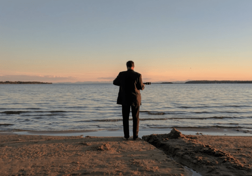 A man in a suit stands on a beach at sunset, playing a guitar by the water's edge.