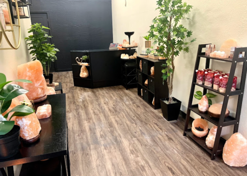 A serene room featuring Himalayan salt lamps, plants, and shelves displaying salt products, with a dark wall backdrop.