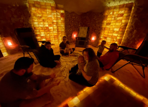 A group of people relax in a softly lit salt room, sitting on the floor with children and lounge chairs around them.