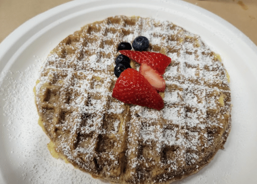 A round waffle topped with powdered sugar and fresh strawberries and blueberries on a white plate.