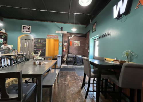 Cozy restaurant interior with wooden tables, teal walls, and a warm atmosphere. A staff member is seen in the background.