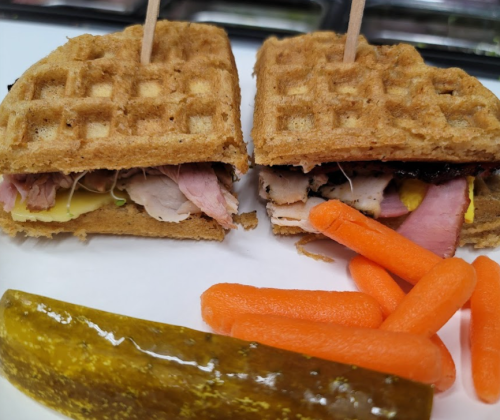 A sandwich made with waffles, filled with meats and mustard, served with baby carrots and a pickle on the side.