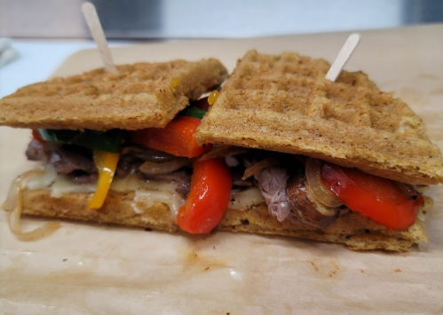 A sandwich made with waffle bread, filled with beef, sautéed peppers, and onions, served on a wooden board.