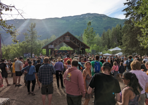 A large crowd gathers at an outdoor music festival, surrounded by trees and mountains under a bright sky.