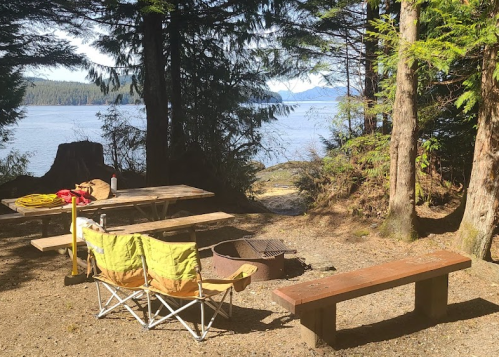 A serene campsite by the water, featuring a picnic table, fire pit, and two yellow chairs surrounded by trees.