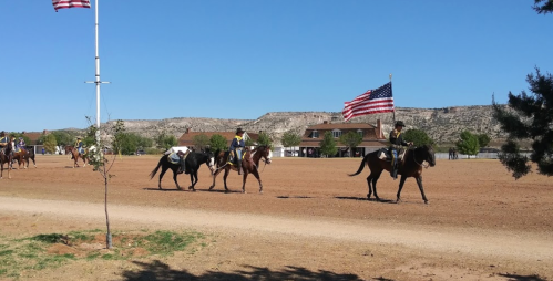 A group of riders on horseback, some carrying American flags, parade across a dusty field with hills in the background.