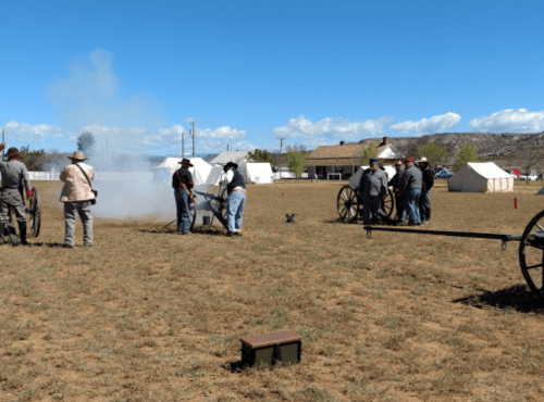 Civil War reenactors firing a cannon in a field, with tents and a blue sky in the background.