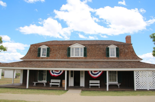 Historic house with a sloped roof, green shutters, and patriotic bunting, set against a blue sky with clouds.