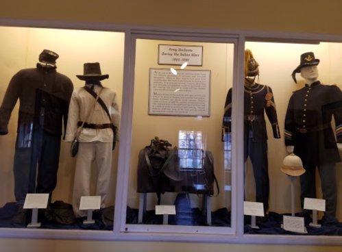 Display case featuring historical military uniforms from 1865-1880, including hats and accessories.