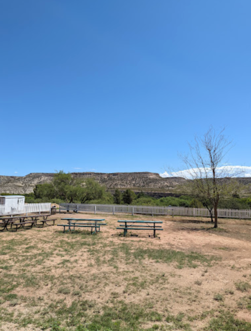 A clear blue sky over a grassy area with picnic tables and distant rocky hills in the background.